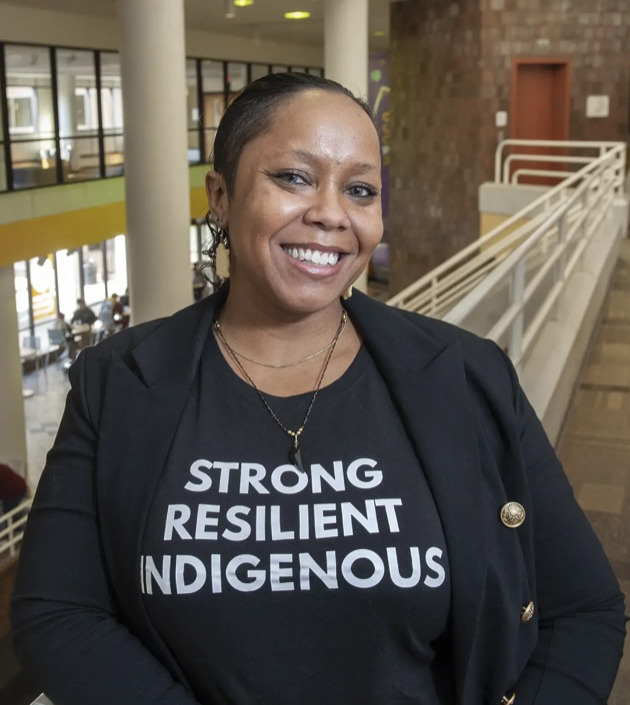 A woman wearing a black blazer with a white graphic T-Shirt that reads: Strong, Resilent, Indigenous
